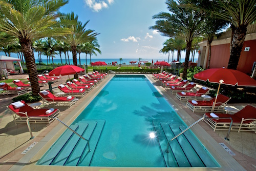 A photo of the beautiful blue pool at Acqualina, set on the beach, surrounded by red lounge chairs and umbrellas.